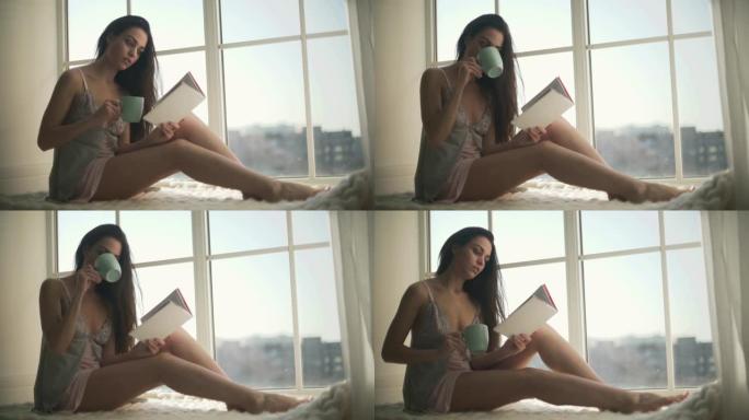 穿着睡衣的漂亮女孩坐在宽阔的窗户旁。穿着睡衣的年轻女子一边喝着晨间咖啡一边看书。