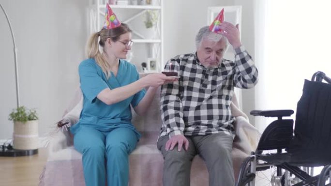 善良的白人妇女在疗养院给白发老人送生日蛋糕。沮丧的退休人员脱下党帽，用手握住头。悲伤，沮丧，衰老。