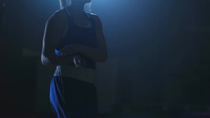 在大厅里训练的年轻美女职业拳击手在蓝色衣服的黑暗房间里，在对立的灯光下跳上绳子