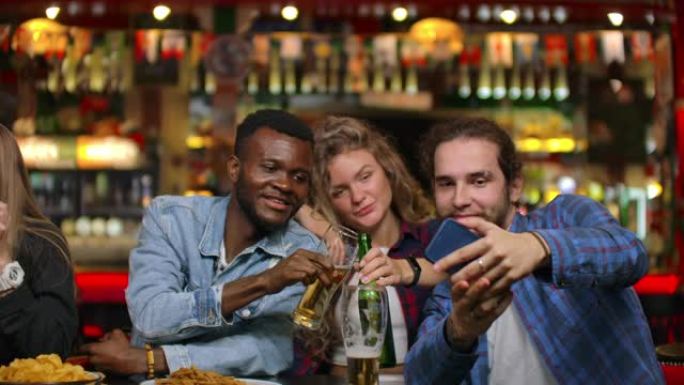 非裔美国人和欧洲朋友坐在酒吧里自拍。一大群朋友坐在一张桌子上的酒吧里聊天，喝啤酒和拍照