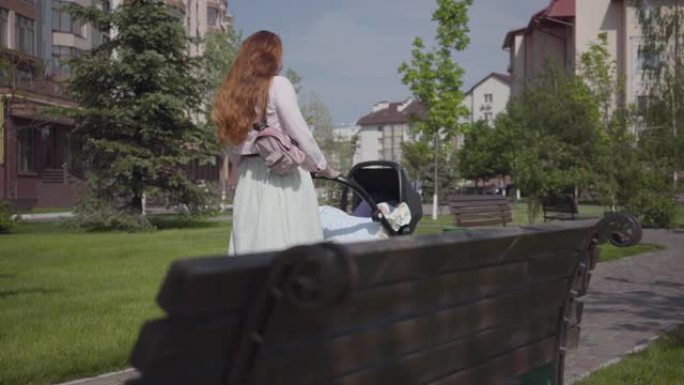 美丽的红发女人走在公园里的一条带婴儿车的石头走道上。