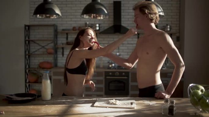 年轻性感性感的女孩在胸罩和英俊的男人在厨房里互相喂辣椒红辣椒。穿着内衣的恩爱夫妻在厨房里嬉戏。缓慢的