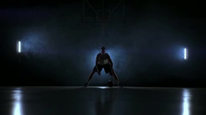 一名男子在黑暗的篮球场上，在烟雾中的篮球环的背景下，展示了由三个灯笼照亮的运球技巧。
