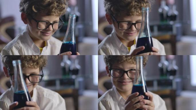快乐的高加索男孩拿着蓝色液体的烧瓶，微笑着。聪明卷发少年实验化学试剂的肖像。电影院4k ProRes