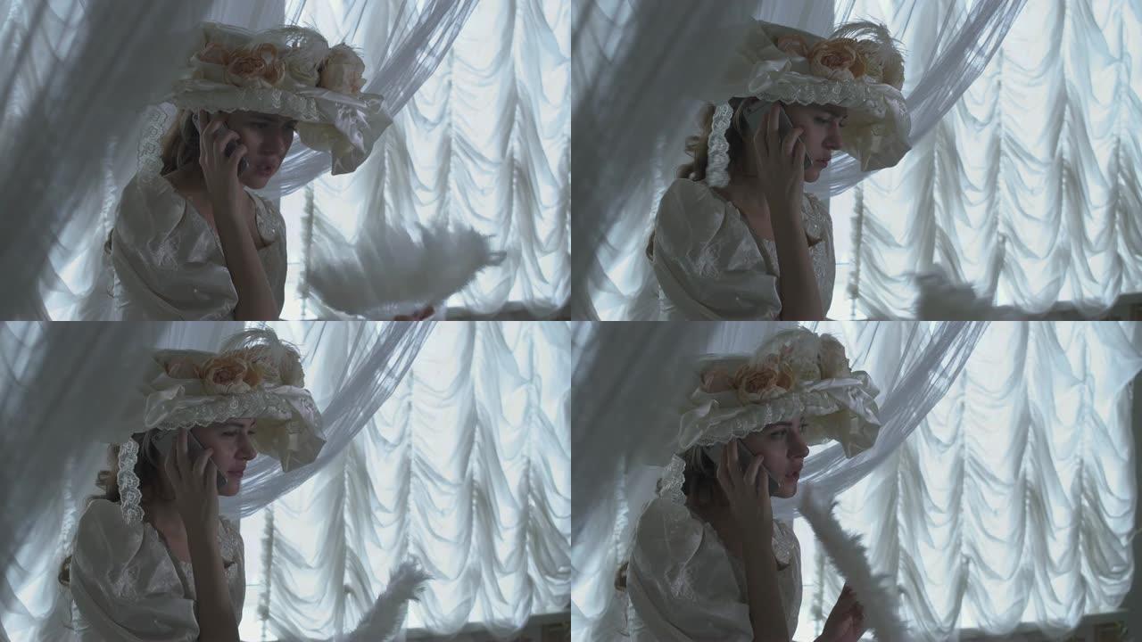 肖像漂亮的年轻微笑的女人，穿着白色复古舞会礼服和白色帽子，站在透明窗帘的背景上，挥舞着风扇在手机上说