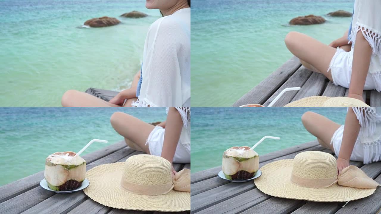 女人戴着一顶海帽她很开心，坐着，椰子在木桥上喝酒，看着海滩海边，云和蓝天是无尽的背景。