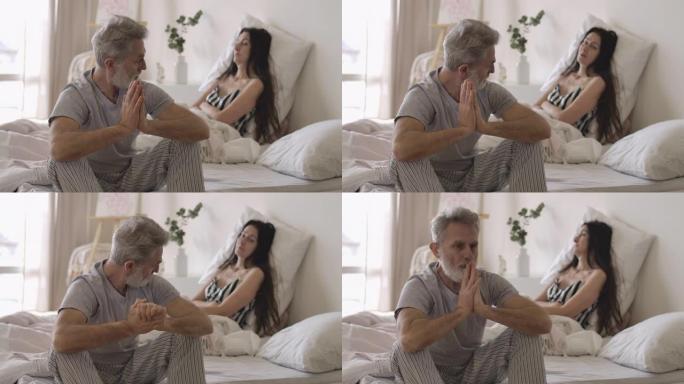 高级英俊的白人男子坐在床边，回头看着躺在背景上的妻子的肖像。早上有感情困难的成熟夫妇。电影院4k P