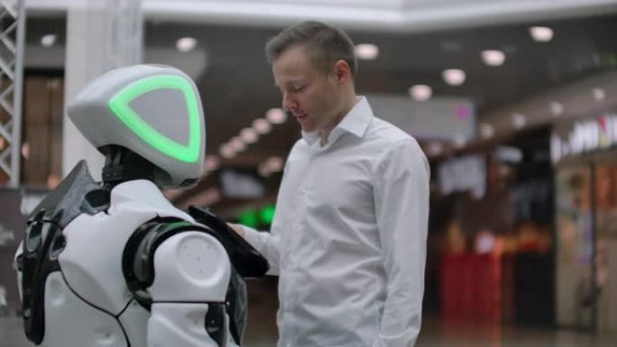 一名男子与机器人机器人站在一起，向他提问，并通过点击机器人身体上的屏幕寻求帮助。