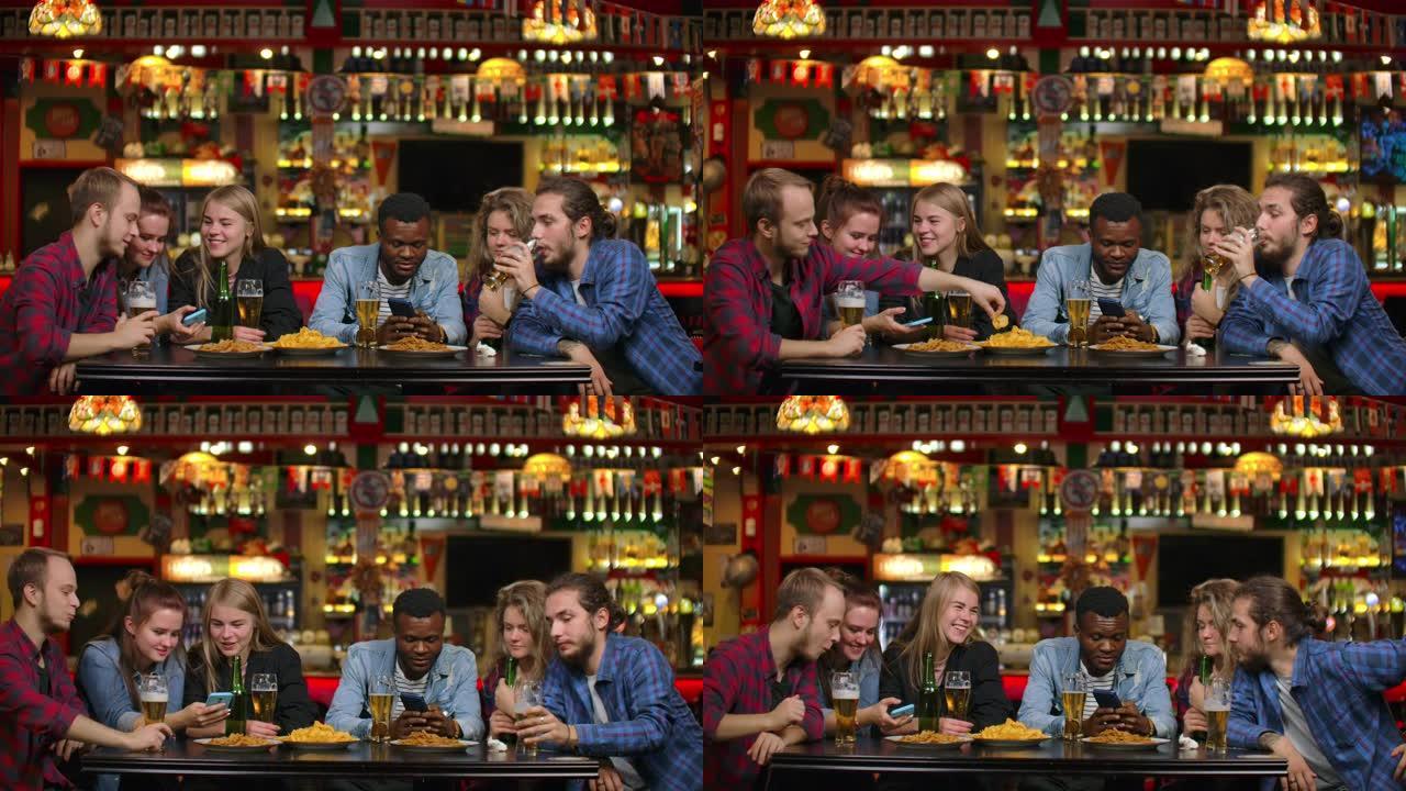 开朗的学生坐在酒吧的桌子旁喝啤酒，吃薯片并在智能手机屏幕上观看照片，讨论照片