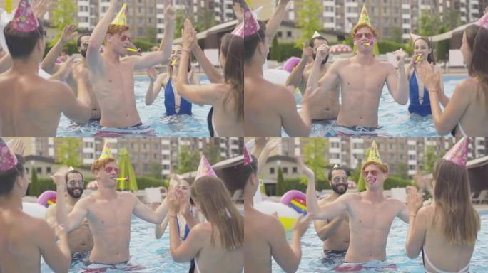 年轻的红发男人和朋友在游泳池里庆祝生日。快乐的年轻高加索人在度假村跳舞的肖像，周围有快乐的人。户外夏