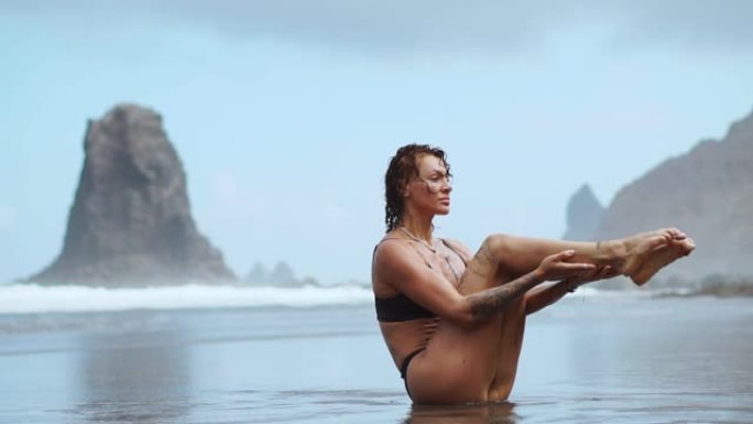 运动女子在沙滩上训练腹肌，黑沙在岩石和波浪的背景下