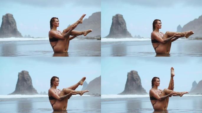 运动女子在沙滩上训练腹肌，黑沙在岩石和波浪的背景下