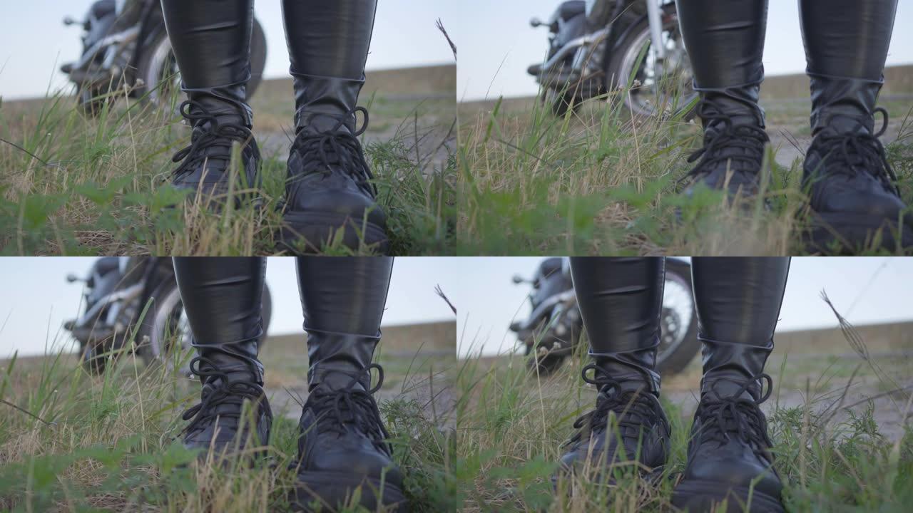 穿着黑色皮裤和运动鞋的不明骑行者的腿站在田野的草地上。