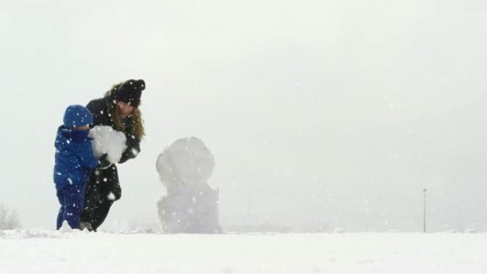 三岁的高加索男孩和他三十多岁的高加索母亲 (都穿着冬装) 在下雪的阴天一起堆雪人
