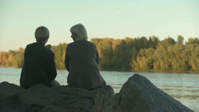 两名老年妇女坐在公园的河岸上扔饭喂鸭子