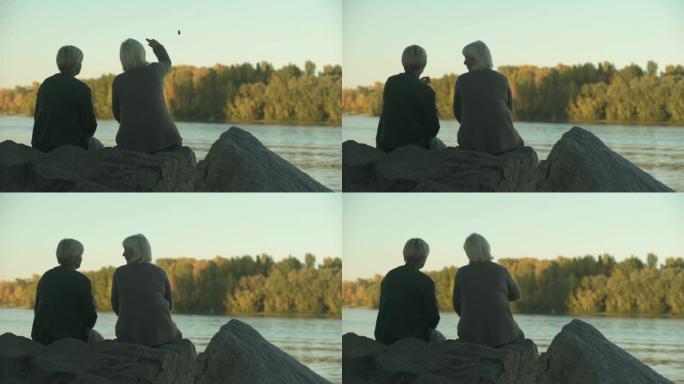 两名老年妇女坐在公园的河岸上扔饭喂鸭子
