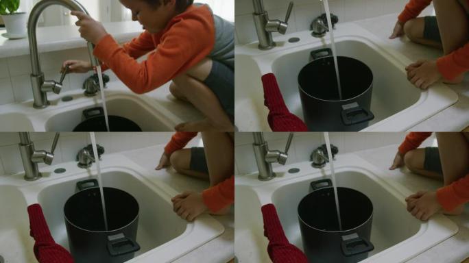 坐在柜台上，一个五岁的白人男孩从厨房水槽里往壶里装满水