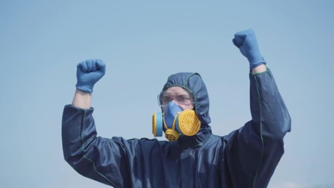 穿着呼吸器和生物危害服的高加索人在天空大喊并表现出力量手势。年轻人厌倦了全球大流行或化学污染，在阳光