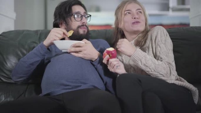 年轻的白人妇女和怀孕的丈夫一起看电视，吃盘子里的薯条。有趣的家庭交换社会角色。开玩笑，有趣，开玩笑。