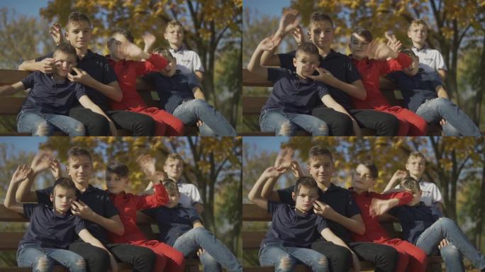 五个男孩坐在长凳上，在相机上挥手。朋友们花时间在户外的大公司里。
