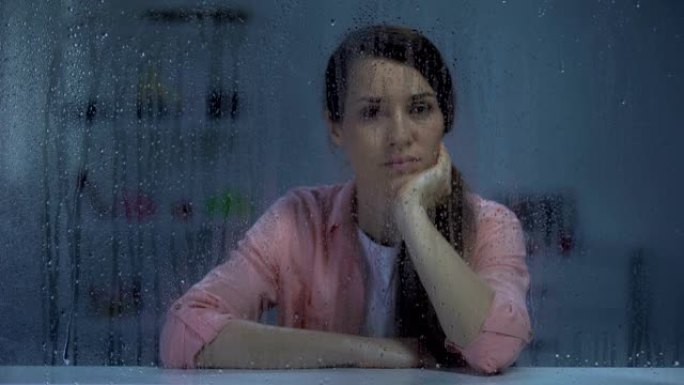 沮丧的年轻女子透过下雨的窗户看着相机，生活问题