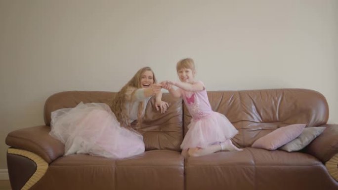 年轻的高加索母亲和她可爱的小女孩穿着同样的裙子跳上皮沙发，打开电视
