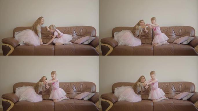 年轻的高加索母亲和她可爱的小女孩穿着同样的裙子跳上皮沙发，打开电视