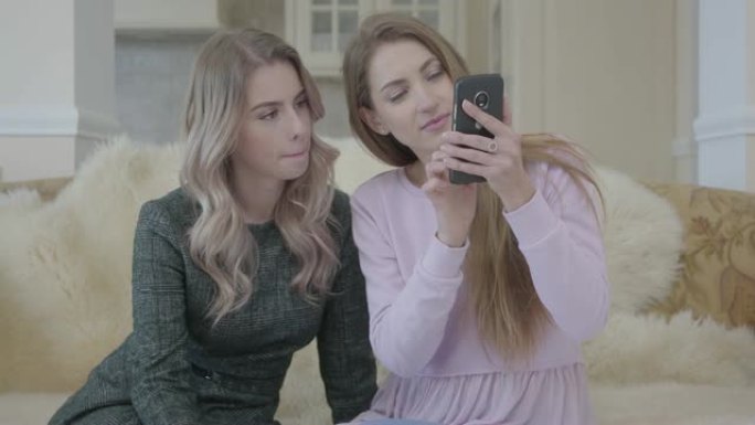 两个穿着时尚连衣裙的漂亮年轻女孩坐在客厅的沙发上看着电话。
