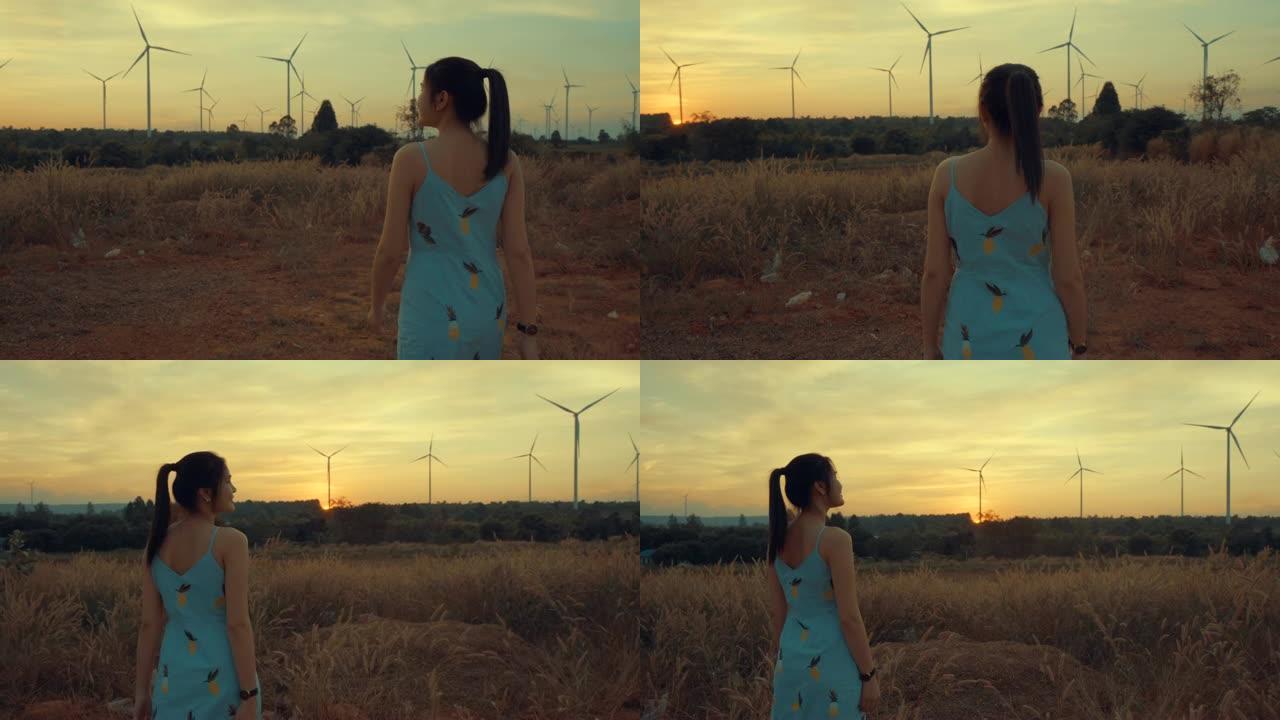 美丽的女孩走在草地上，她微笑着暮色背景是一台风力涡轮机。