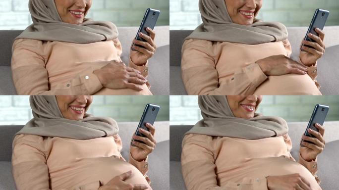 快乐的穆斯林孕妇在智能手机上观看卡通并微笑，应用程序