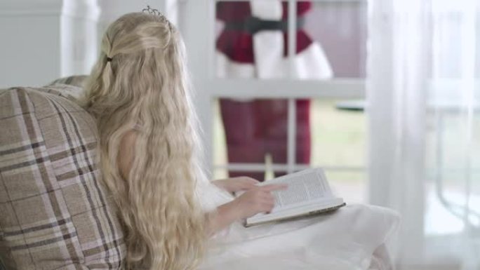 漂亮的白人金发女孩坐在沙发上看书，圣诞老人站在玻璃入口门口敲门。儿童合上书和挥手。电影院4k Pro