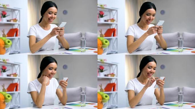 迷人的微笑女性通过智能手机应用程序聊天，坐在家里的桌子上