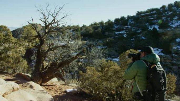 一位四十多岁的白人男性摄影师穿着远足装备，冬天在科罗拉多州西部的高沙漠中拍摄了一棵枯树的照片 (科罗