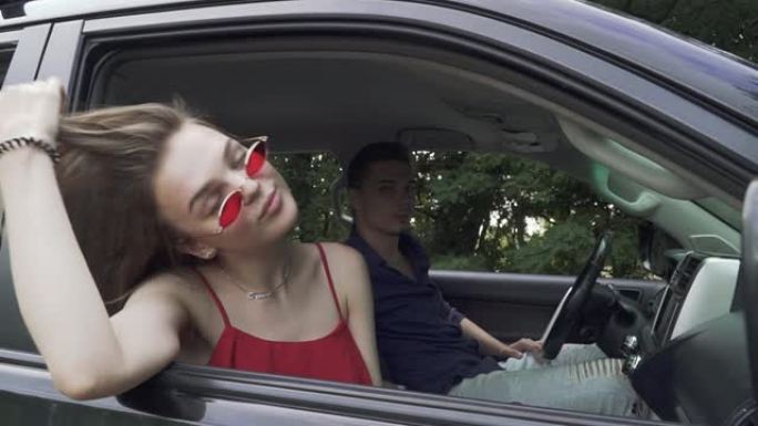 年轻夫妇开车旅行。穿着红色衬衫和太阳镜的女士露出舌头。人在开车。男人和女人在一起。乘坐现代汽车旅行