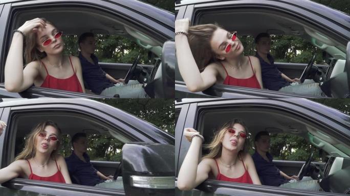 年轻夫妇开车旅行。穿着红色衬衫和太阳镜的女士露出舌头。人在开车。男人和女人在一起。乘坐现代汽车旅行