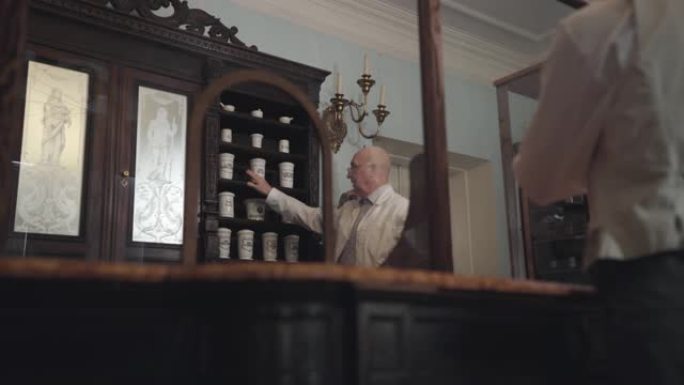 老明智的高加索药剂师在眼镜上向顾客展示装有药品的老式瓶子。19世纪药店，复古，复古，制药博物馆。