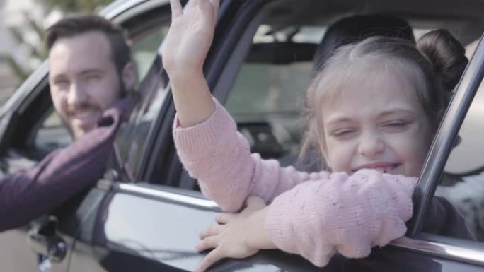 坐在汽车后座上的小女孩挥舞着手，近距离地看着镜头。