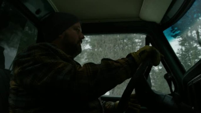 一个三十多岁的白种人留着胡须，在一个下雪的冬天，他在常绿的森林里开着卡车。