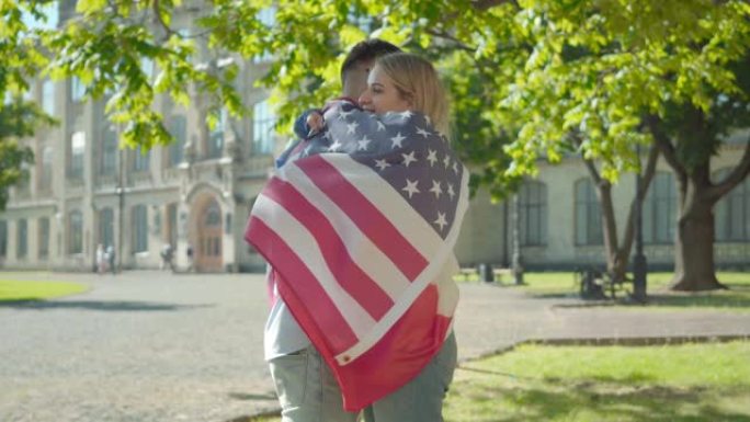 充满爱心的国际学生在阳光明媚的大学院子里拥抱的侧视图。正面的美国男人和法国女人被国旗包裹的肖像。教育
