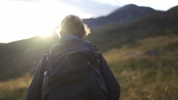 日落时分，男性徒步旅行者带着背包在山顶下穿过高高的草丛