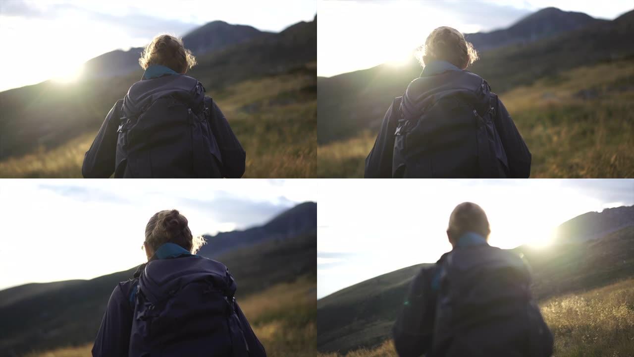日落时分，男性徒步旅行者带着背包在山顶下穿过高高的草丛