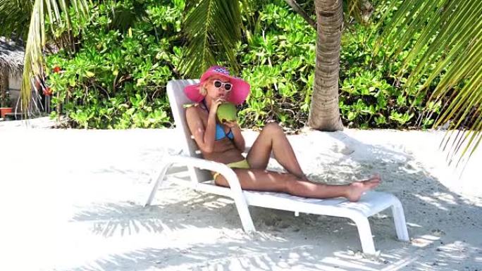 时髦的女人坐在棕榈树下，在马尔代夫喝椰子汁