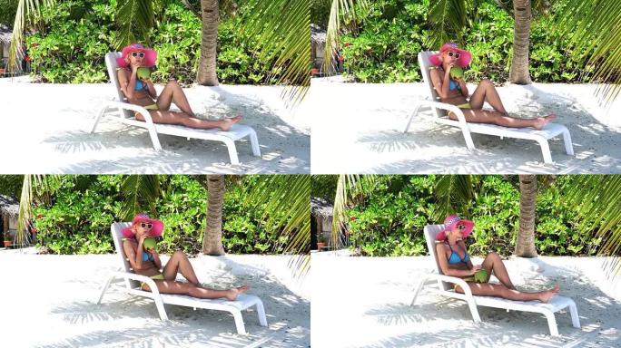 时髦的女人坐在棕榈树下，在马尔代夫喝椰子汁