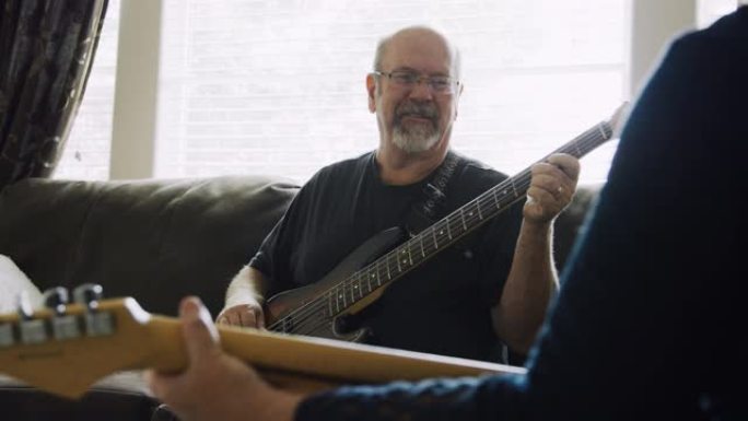 一名五十多岁的高加索人白天在家庭客厅与另一个人演奏他的五弦电贝斯吉他