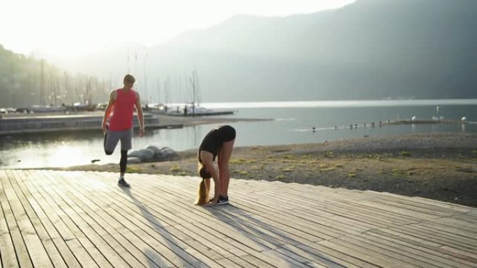 年轻的运动夫妇在日出时沿着山下的湖岸伸展