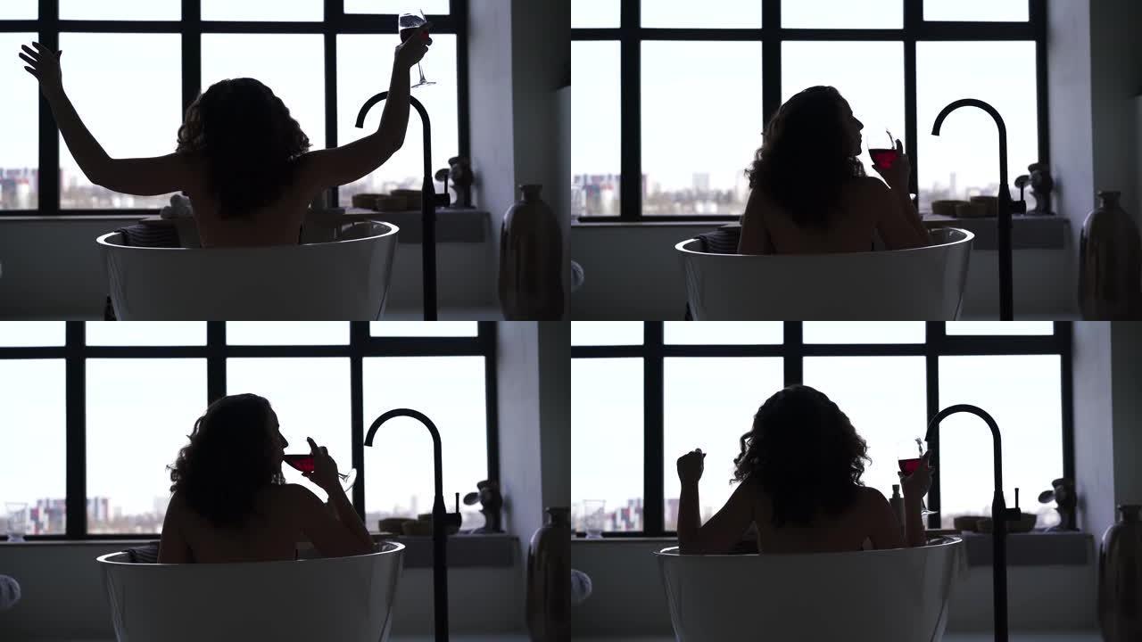 年轻女子在浴缸里喝葡萄酒的剪影。晚上，开朗的高加索女孩在大窗户前的浴室里休息。后视图。