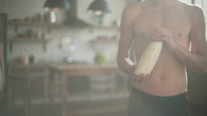 裸体的男人拿着一瓶牛奶站在阳光下的厨房里。穿着睡衣的可爱女孩来到那个男人，拿着牛奶和树叶。
