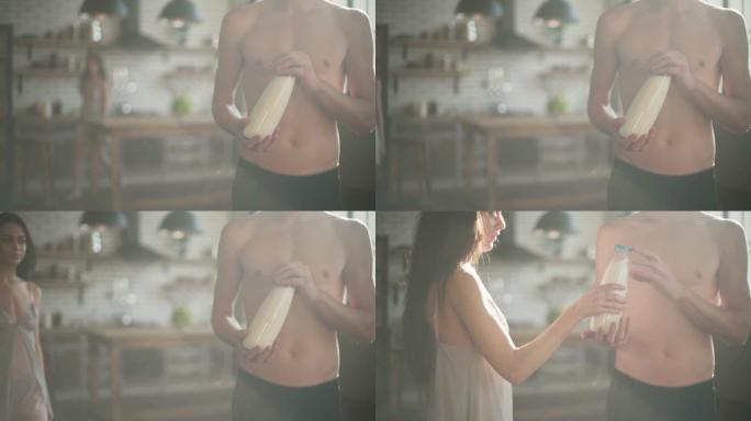 裸体的男人拿着一瓶牛奶站在阳光下的厨房里。穿着睡衣的可爱女孩来到那个男人，拿着牛奶和树叶。