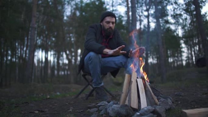 一个年轻的大胡子家伙坐在椅子上，在火旁热身。森林里的火堆。