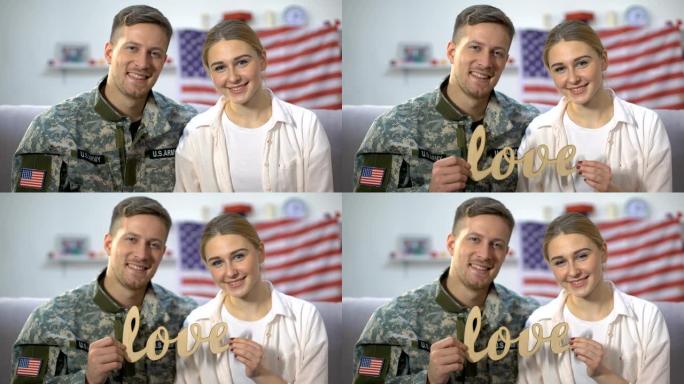 微笑的美国士兵和他的女朋友在相机上展示木制的爱的标志